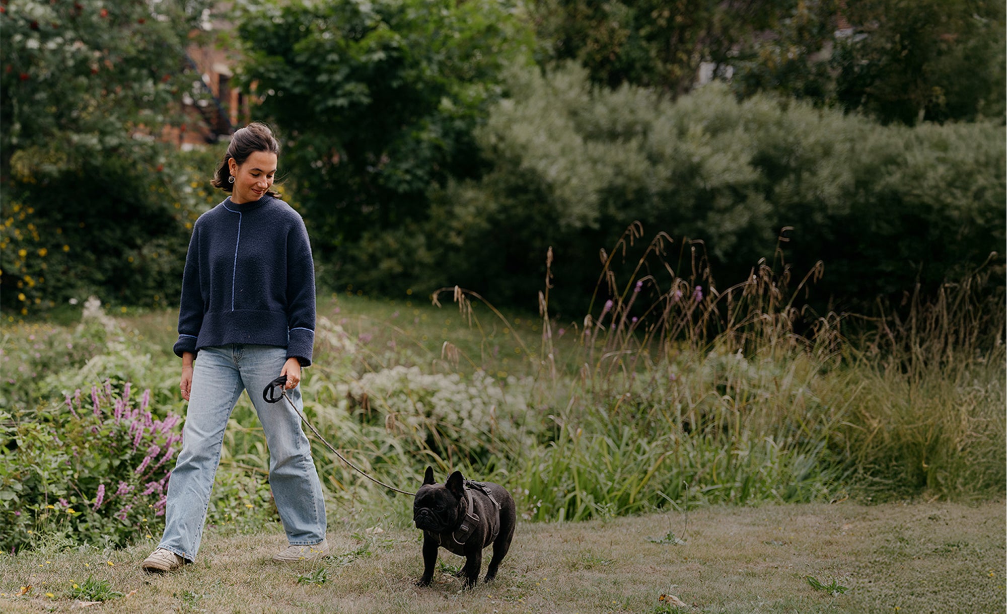 Person walking a small black dog in a natural setting with greenery and flowers.