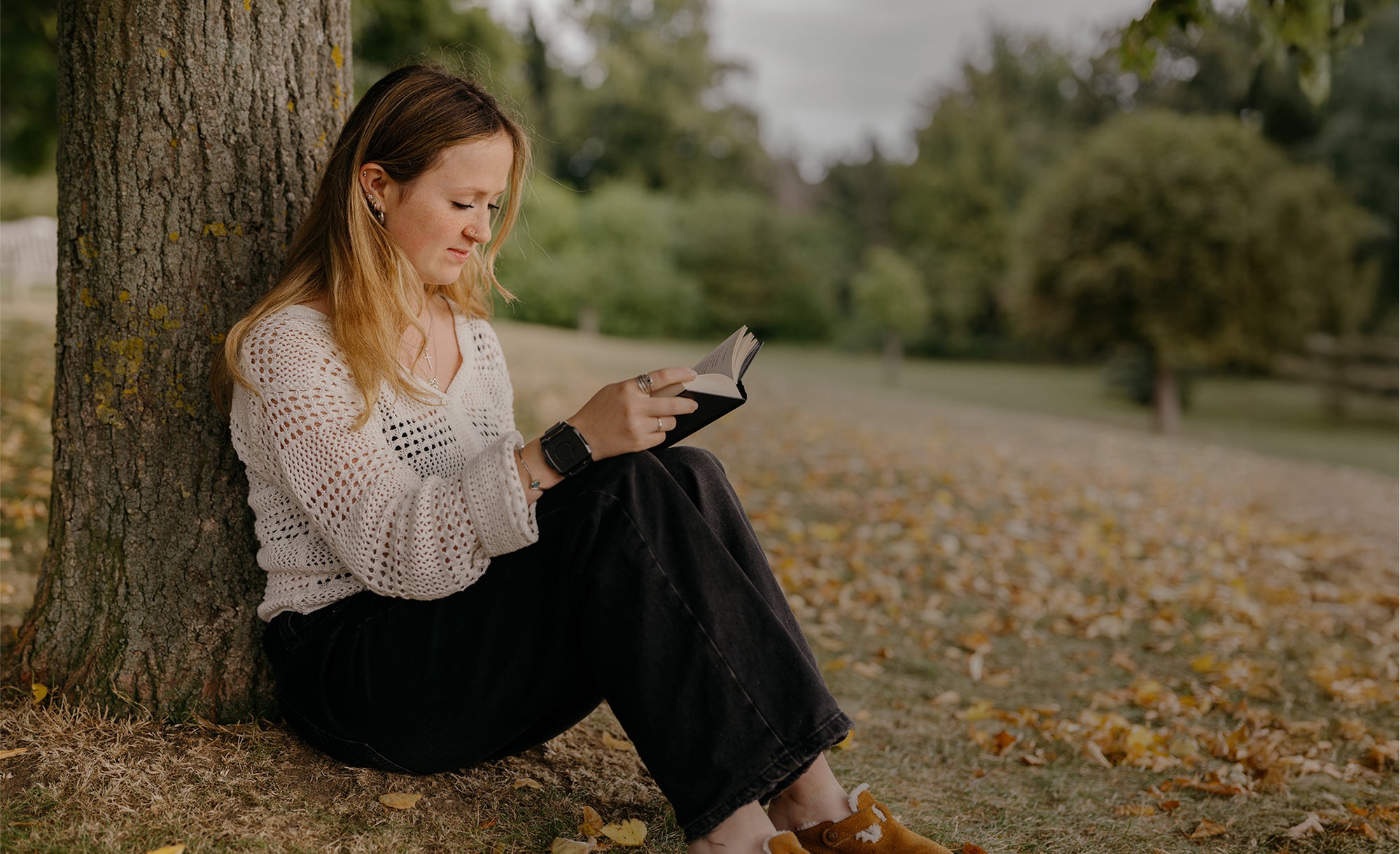 Woman reading a book by a tree in a park with fallen leaves on the ground.